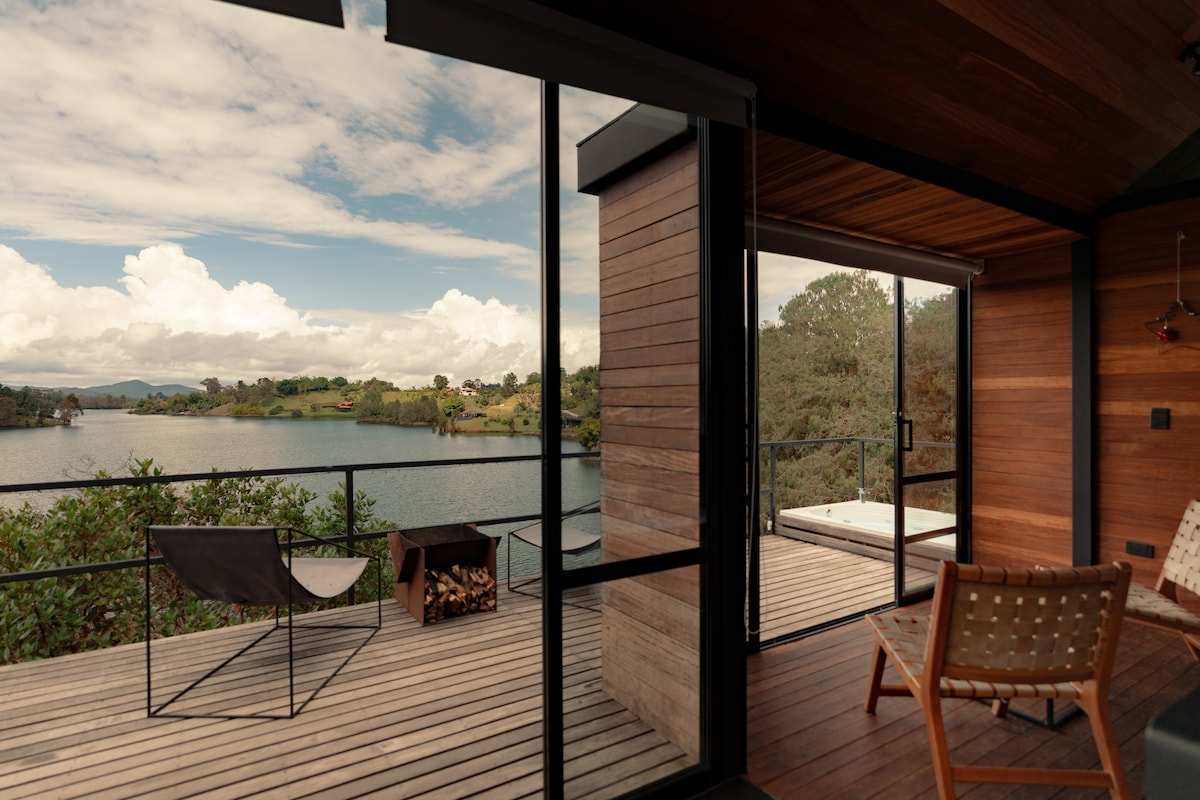 A spacious balcony features panoramic views of the lake and surrounding hills, furnished with a cozy chair and a simple table. Large glass doors open to an inviting interior space, enhanced by natural wood finishes and ample sunlight.