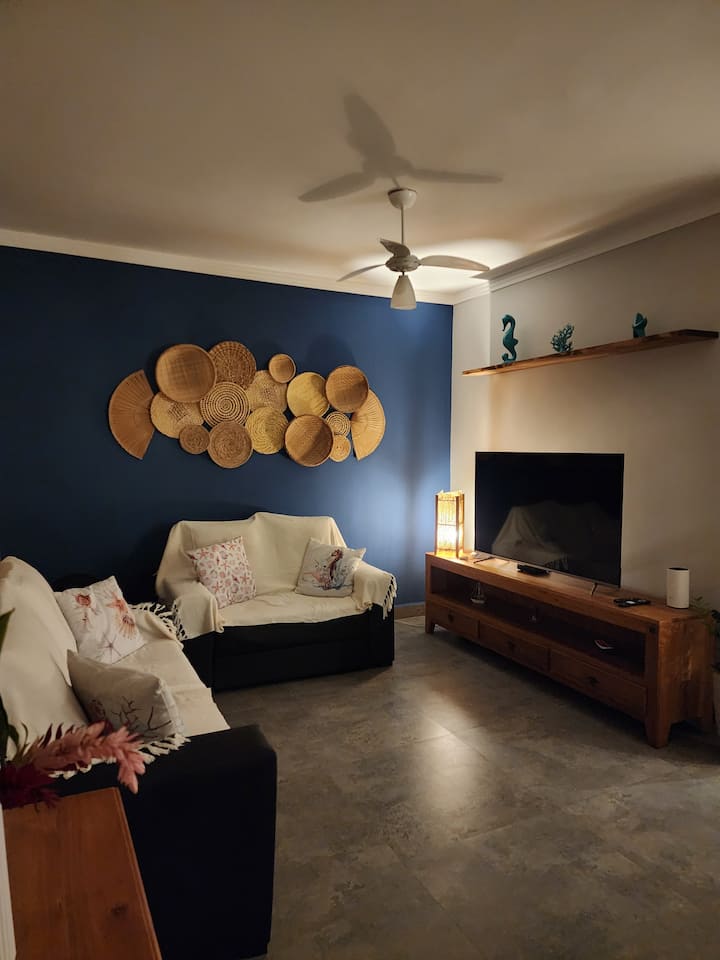 Salon avec deux canapés en cuir, des couvertures en lin, des oreillers et des décorations avec des arrangements de paille et des objets nautiques. Sur l'étagère, une lampe et un téléviseur de 50 po, un routeur Wi-Fi, un ventilateur de plafond