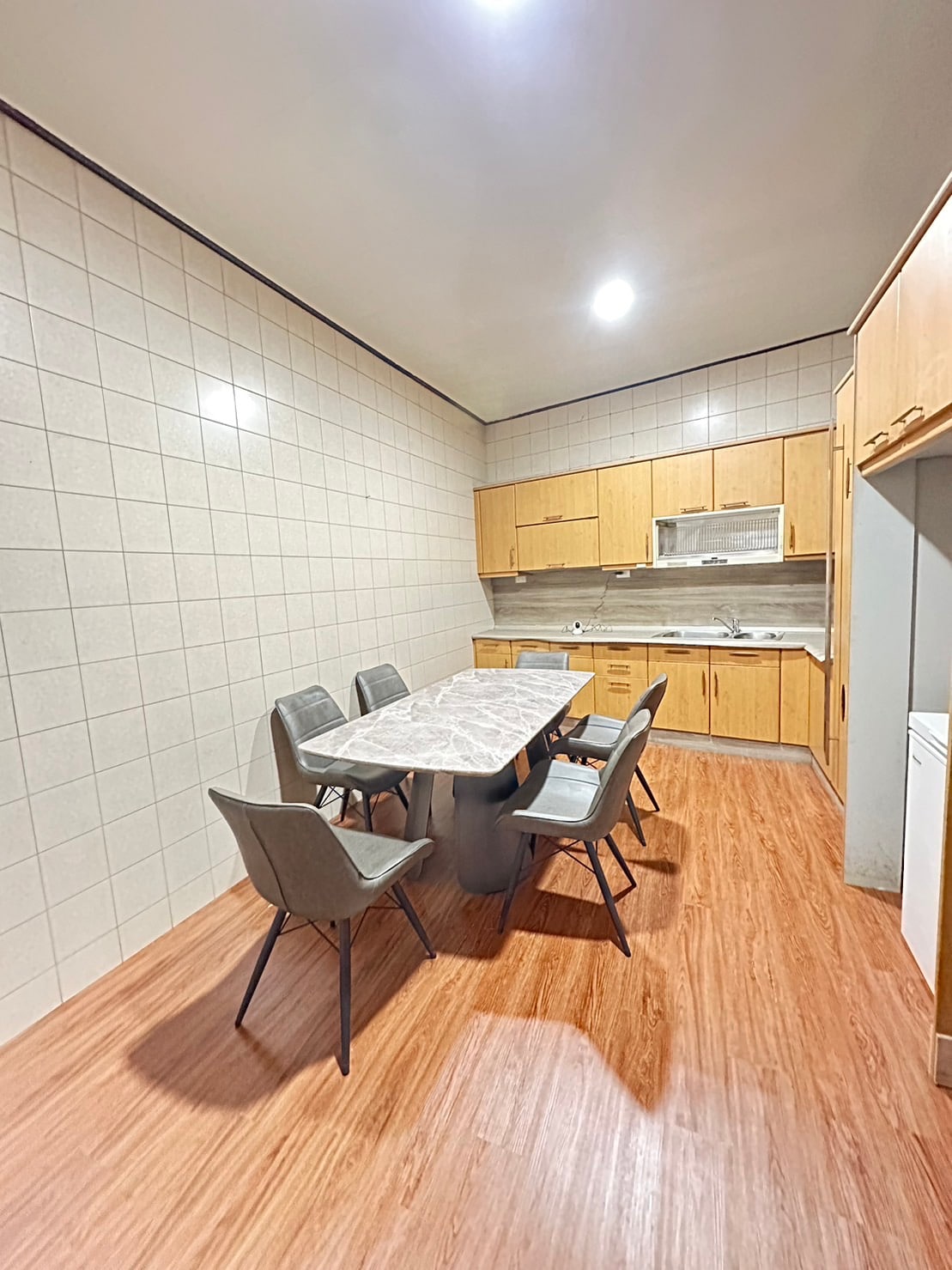 A bright kitchen area features a marble-topped dining table surrounded by six modern chairs. Wooden cabinets and a countertop provide ample storage and workspace. The warm-toned flooring complements the light-colored tiles, creating a functional and inviting cooking space.