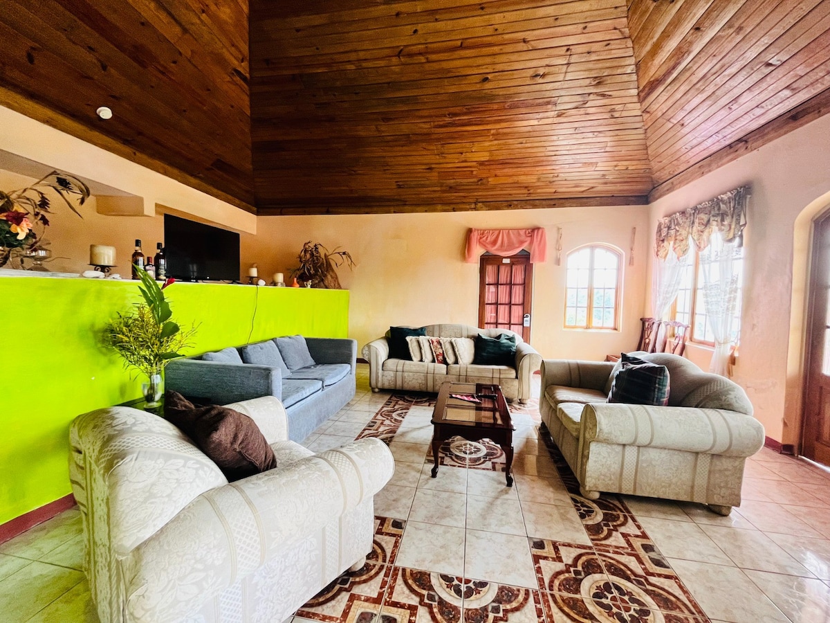 A spacious living area features high wooden ceilings and a mix of comfortable seating options arranged around a central coffee table. Soft lighting enters through multiple windows, complemented by vibrant wall colors and decorative elements that create an inviting ambiance.