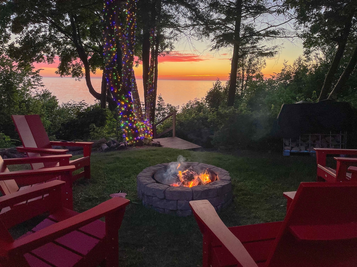 A tranquil fire pit area features red Adirondack chairs arranged around a stone ring, with flames dancing inside. In the background, colorful lights adorn the trees, and the sunset paints the sky above Lake Huron, creating a serene atmosphere.