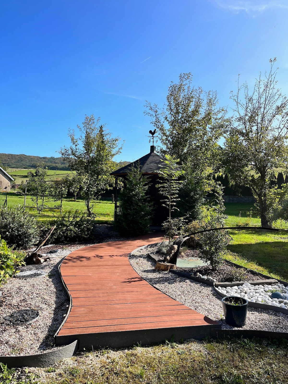 A winding pathway made of warm-toned wood leads through a landscaped garden, surrounded by lush greenery and decorative stones. A small gazebo is positioned in the distance, complementing the serene atmosphere of the outdoor space under a clear blue sky.