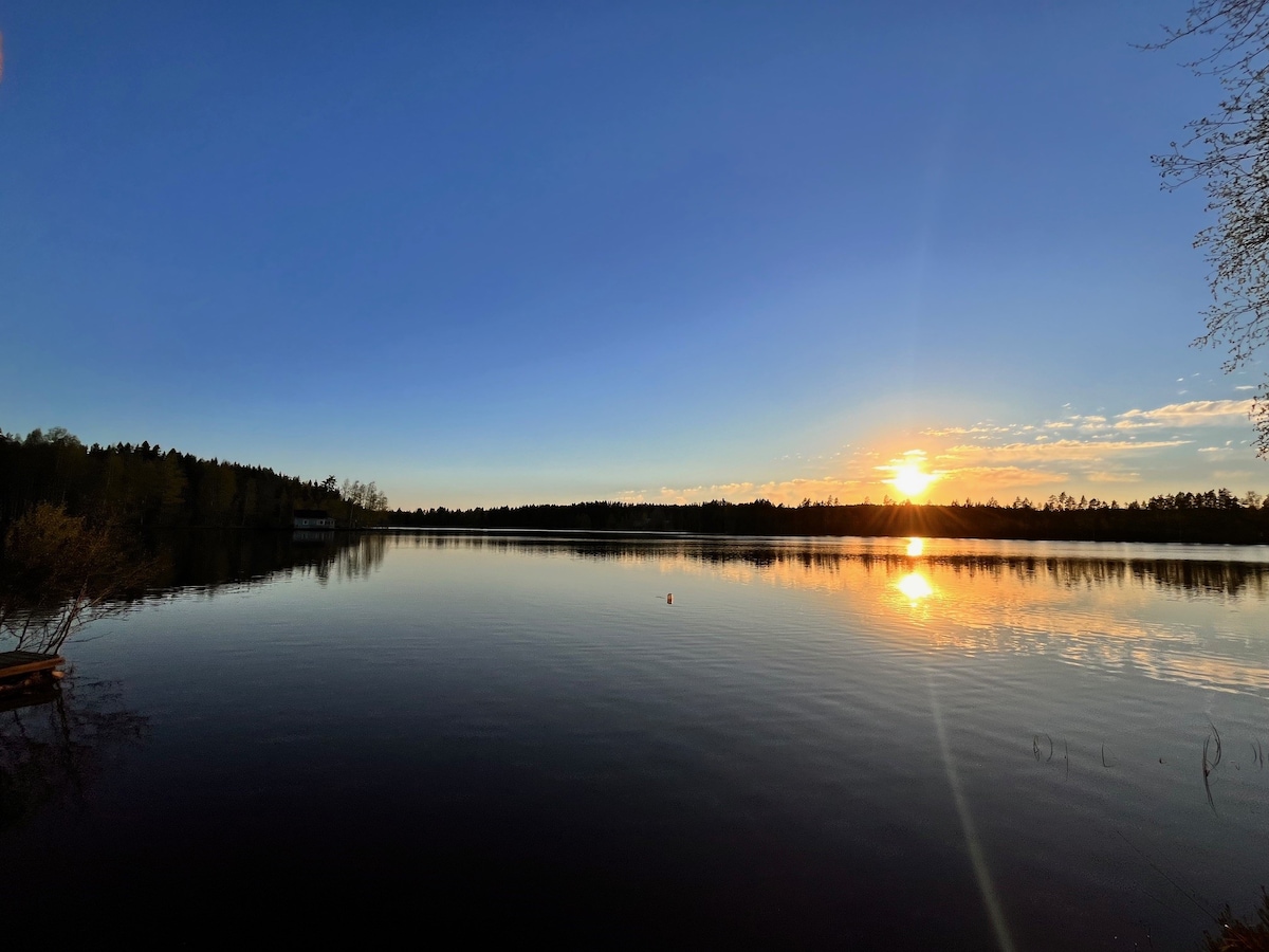 A serene lake is captured at sunset, with the sun reflecting on the calm water. Surrounding forests create a peaceful backdrop, while soft clouds float in the sky. The scene suggests a quiet atmosphere, inviting relaxation and appreciation of nature.