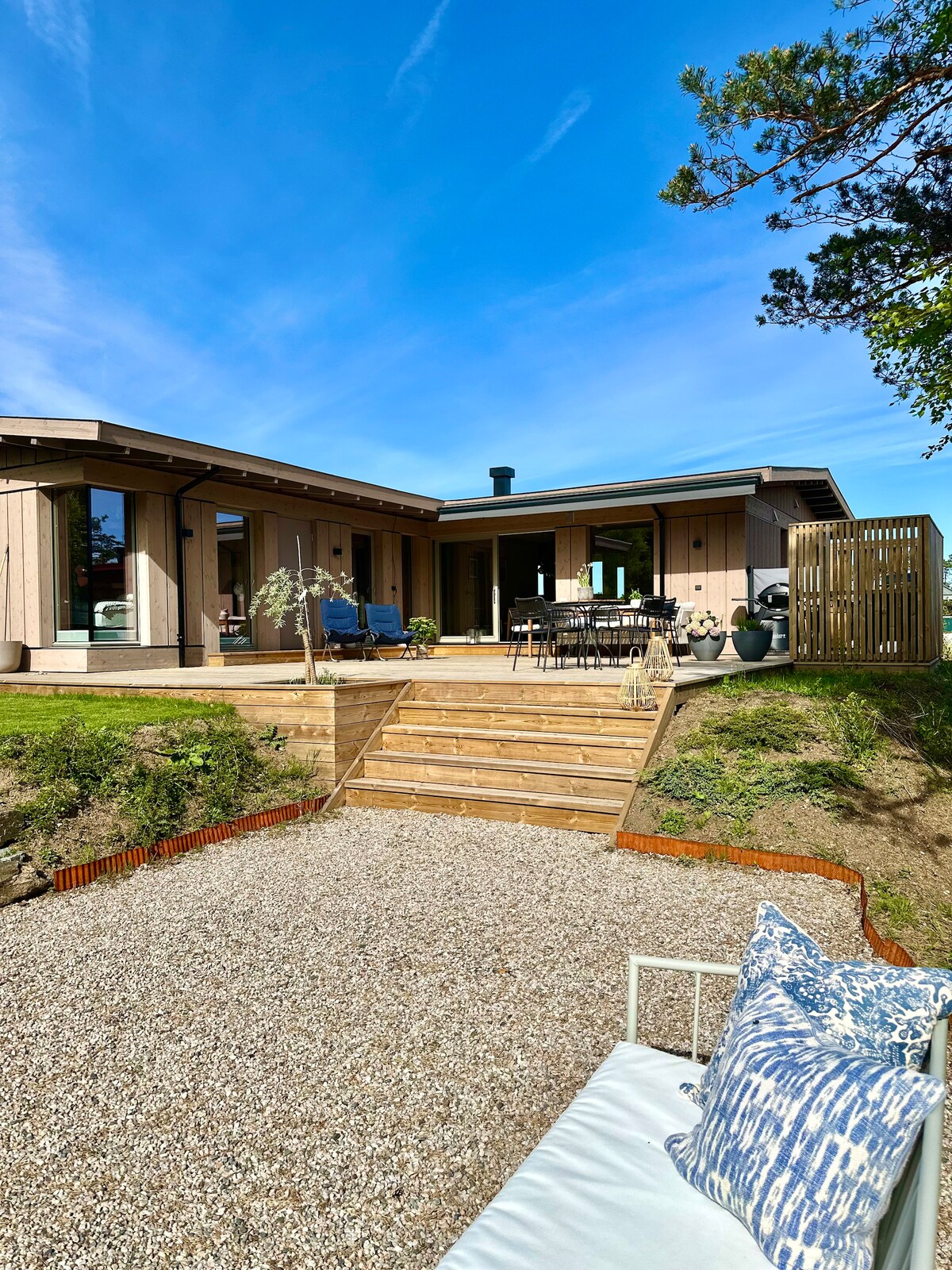 The exterior of the newly built cabin features contemporary design elements with large windows and a welcoming entrance. Wooden steps lead to a spacious terrace with outdoor seating and a dining area, surrounded by grass and decorative landscaping.
