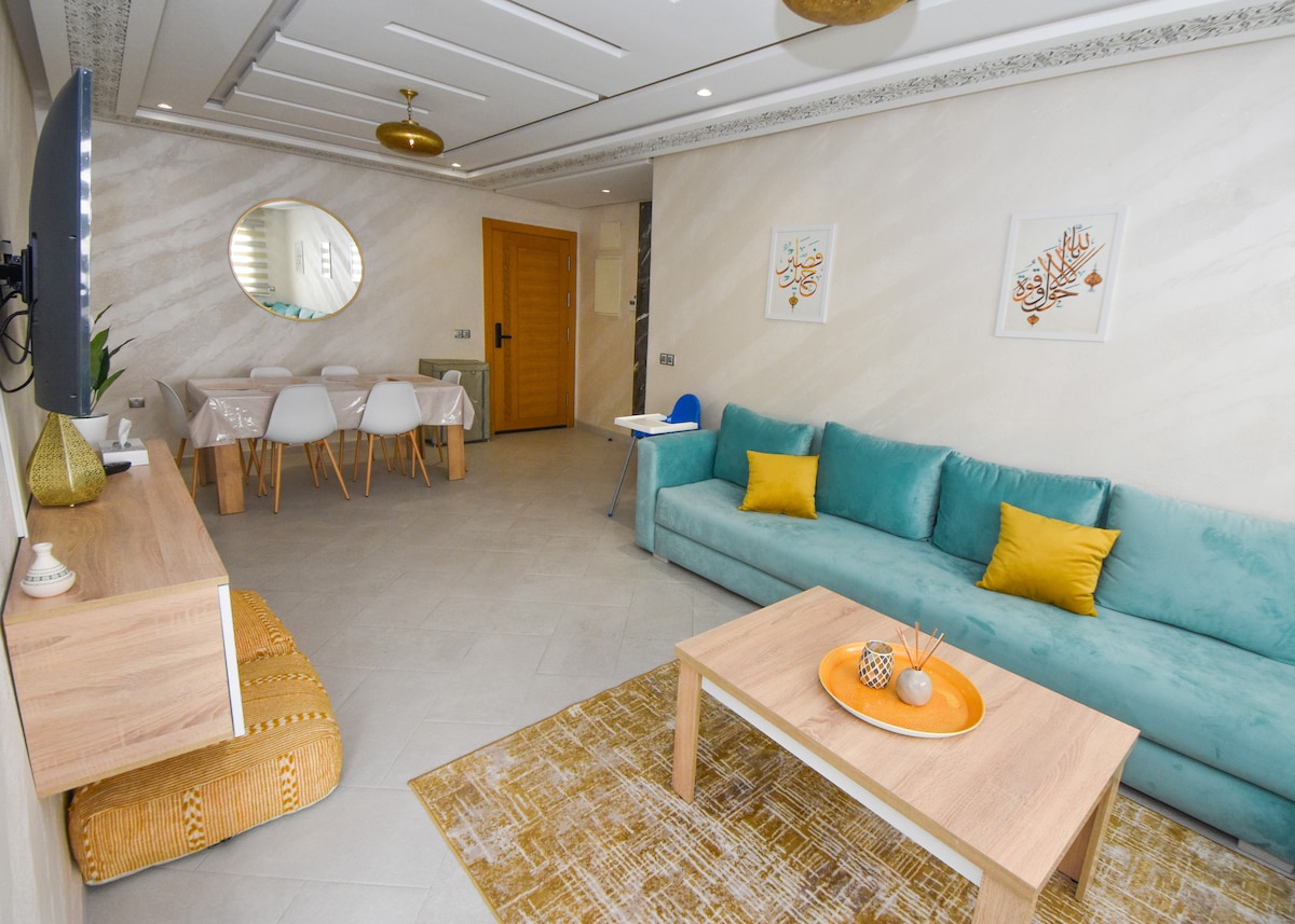 A modern living area features a soft teal sectional sofa with accent yellow cushions. A wooden coffee table is set in front, adorned with decorative items. A dining table with white chairs is visible in the background, surrounded by light-colored walls and artwork.
