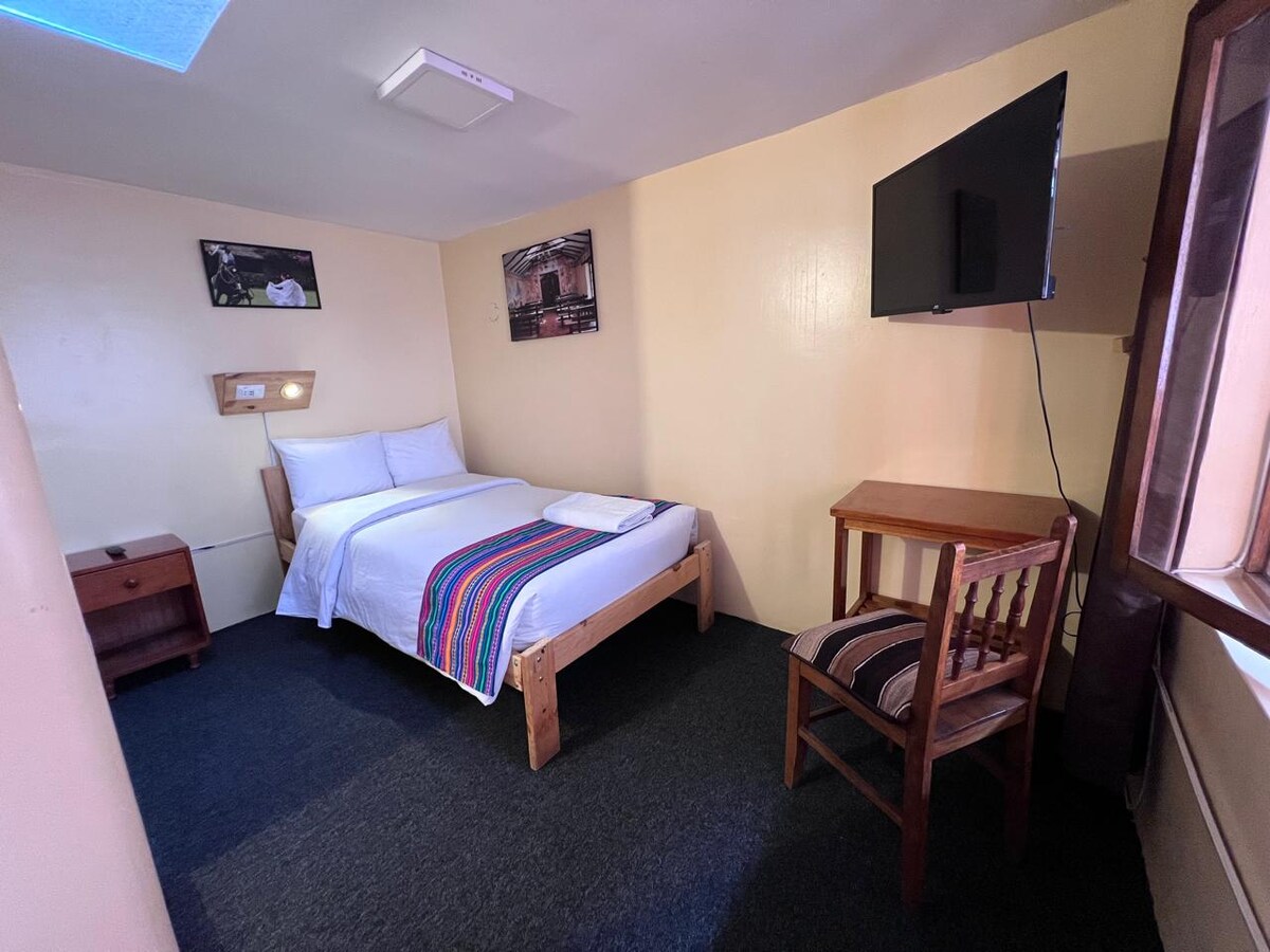 A well-lit room features a spacious bed adorned with a colorful striped blanket. Natural light floods in through the window, which complements a simple wooden chair and bedside table. A wall-mounted TV is positioned across from the bed, enhancing the room's functionality.