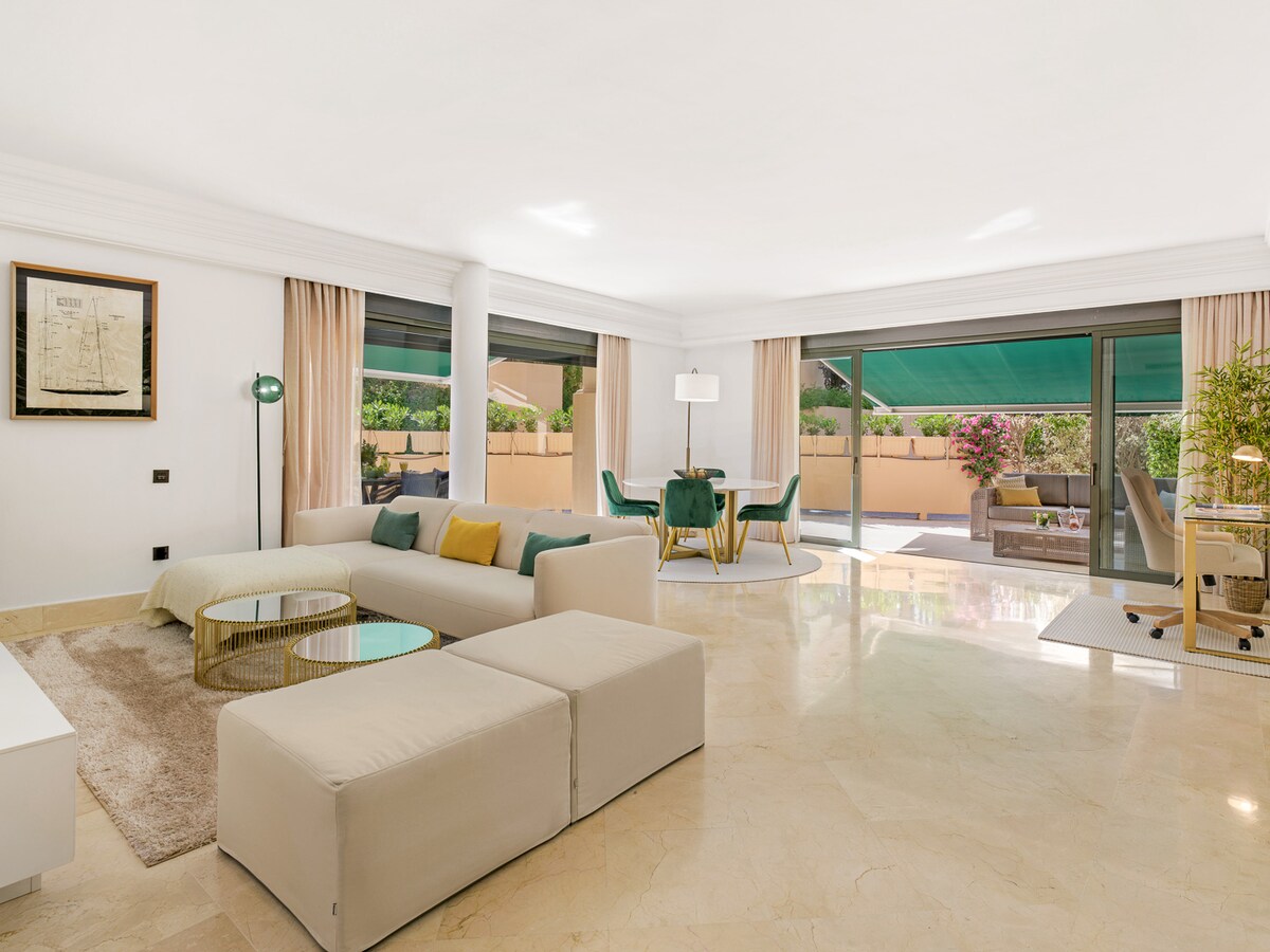 A spacious living area is showcased, featuring a light-colored sectional sofa and a glass coffee table. Large sliding doors provide access to the outdoor terrace and gardens, allowing natural light to fill the room. Neutral tones create a soothing atmosphere.