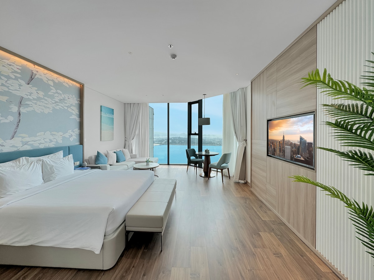 A contemporary room is illuminated by natural light streaming through floor-to-ceiling windows, revealing expansive views of Ha Long Bay. A spacious bed is complemented by a seating area and a smart TV positioned on a wooden wall. A dining table for four is nearby.