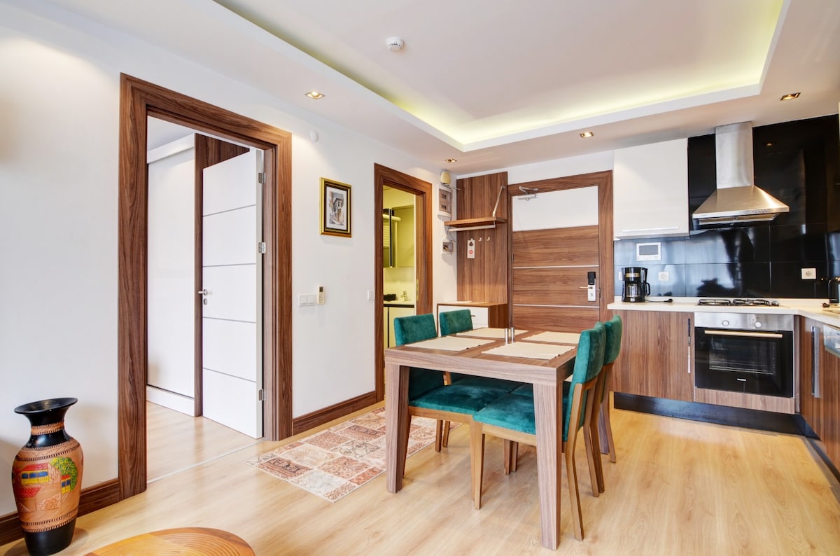 A dining area with a wooden table surrounded by four green chairs is featured. The space showcases modern kitchen amenities, including a stove and coffee maker. Light fixtures create a subtle glow while contrasting textures from the flooring and cabinetry enhance the contemporary aesthetic.