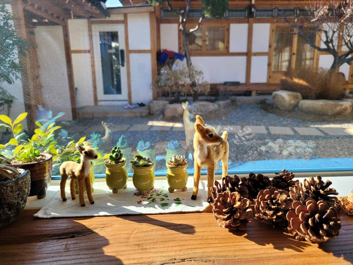 A wooden table displays small ornamental deer figures alongside potted plants. The background features a courtyard with traditional architecture, showcasing natural stones and greenery. Pinecones are arranged on the table, enhancing the rustic appeal of the space.