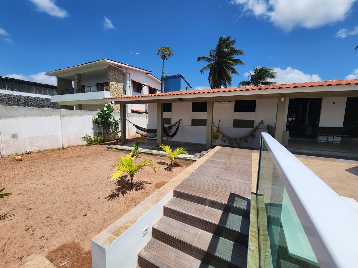 A spacious outdoor area features a light-colored tile patio leading to a well-kept garden with small plants. Multiple hammocks are hung for relaxation, while the background reveals a two-story structure with a textured stone facade and lush palm trees under a bright blue sky.