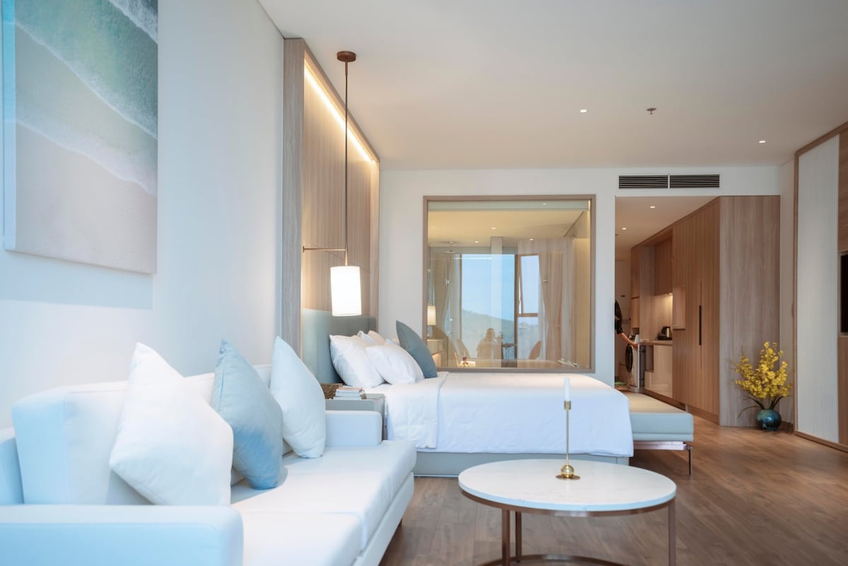 A modern living space is featured with a plush white sofa adorned with light blue pillows. A round coffee table sits in front, while a large bed is visible in the background. A spacious kitchenette can be seen through glass doors, complementing the open and airy design.