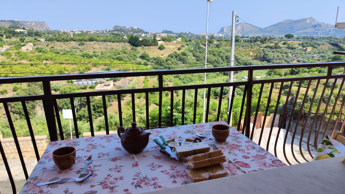 A table set on a balcony features a floral tablecloth with pottery cups and a teapot. The view displays lush green hills under a clear sky, creating a serene backdrop. The railing adds depth to the scene, framing the outdoor setting.