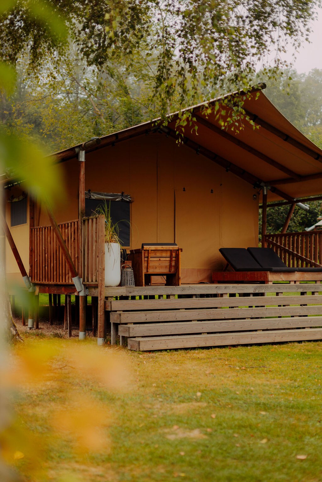 The exterior of a nature lodge is visible, featuring a solid wooden deck with two sun loungers positioned for relaxation. A prominent wooden railing surrounds the entrance, and a decorative pot is placed nearby. The soft, earthy tones of the building blend harmoniously with the surrounding greenery.