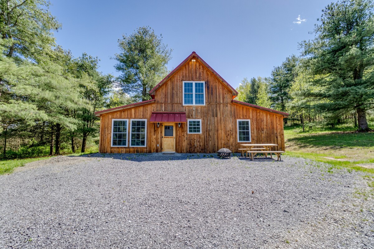 A contemporary cabin features a wooden exterior with a sloped roof and large windows that allow natural light. Gravel serves as the driveway, while a picnic table is positioned in front of the cabin, surrounded by tall pine trees.