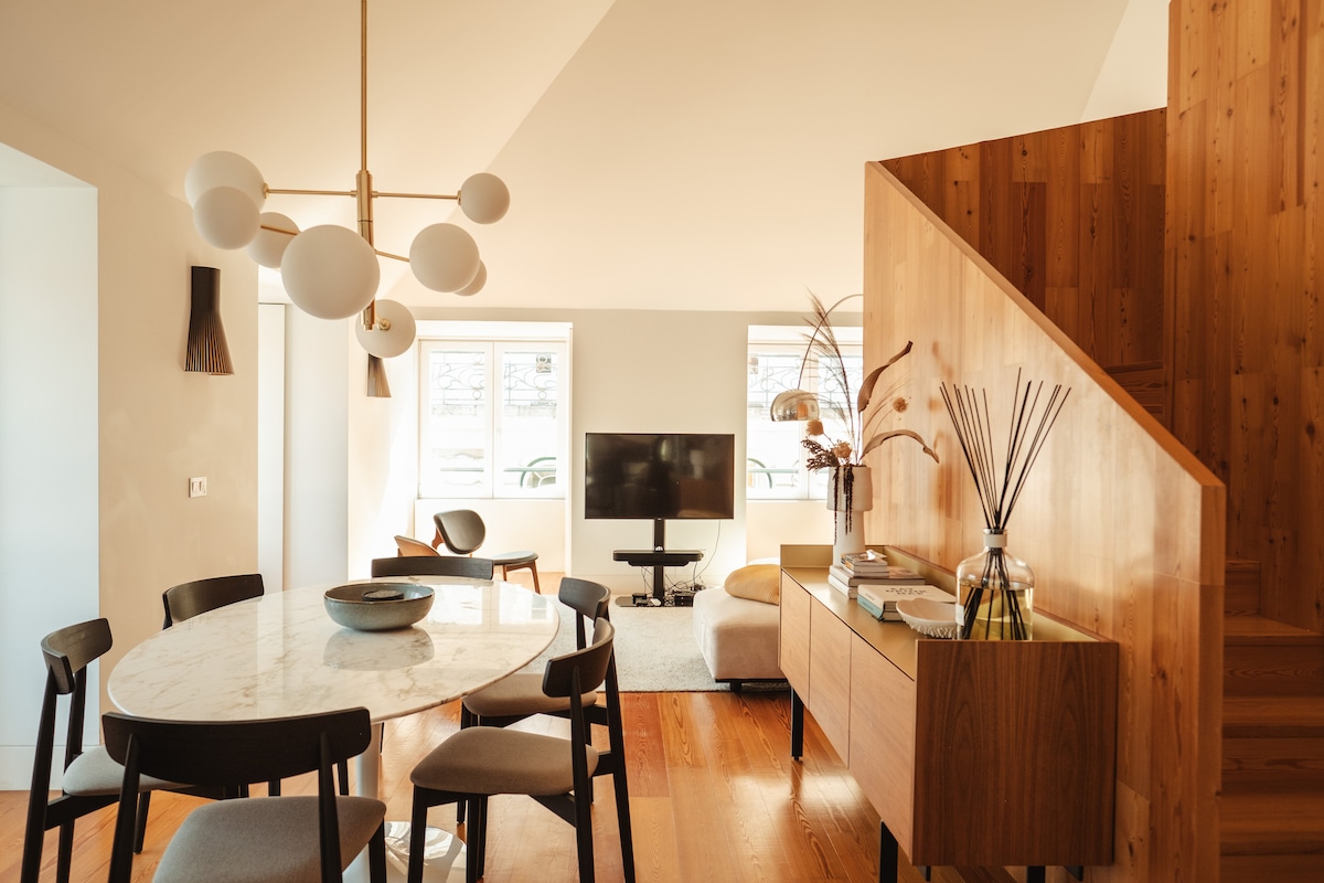 The light-filled living and dining area features a round marble dining table surrounded by dark chairs. A staircase leads to the upper floor, while a comfortable seating area includes a flat-screen TV and decorative elements on a wooden sideboard.