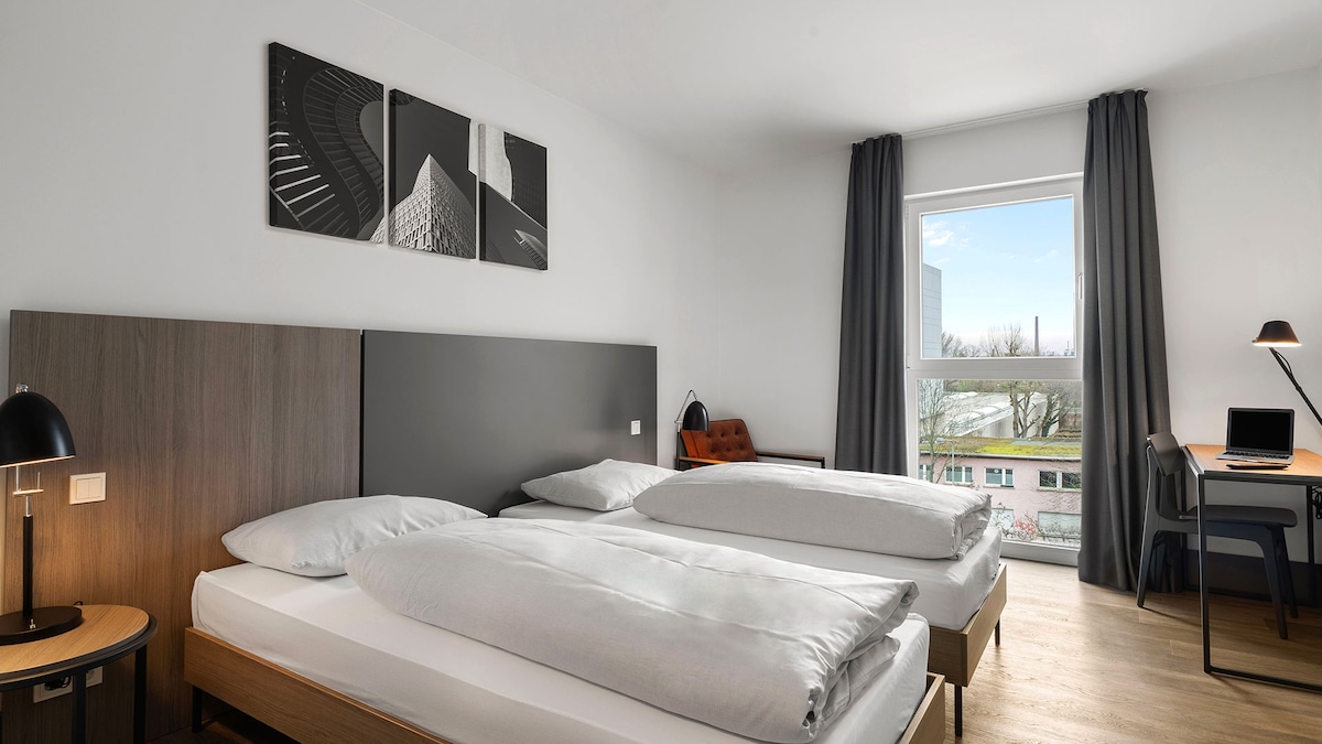 A bright and airy guest room features two spacious beds with crisp white linens topped with soft comforters. A large window allows natural light to fill the room, complemented by light grey drapes. A small desk with a lamp and a comfortable chair can be seen.