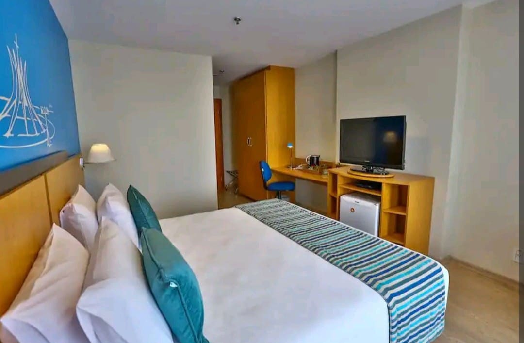 A modern hotel room is showcased, featuring a spacious bed with green and white accent pillows. A wooden desk and chair are visible along with a television mounted on the wall. A compact refrigerator sits beneath the desk, contributing to the room's functional layout.