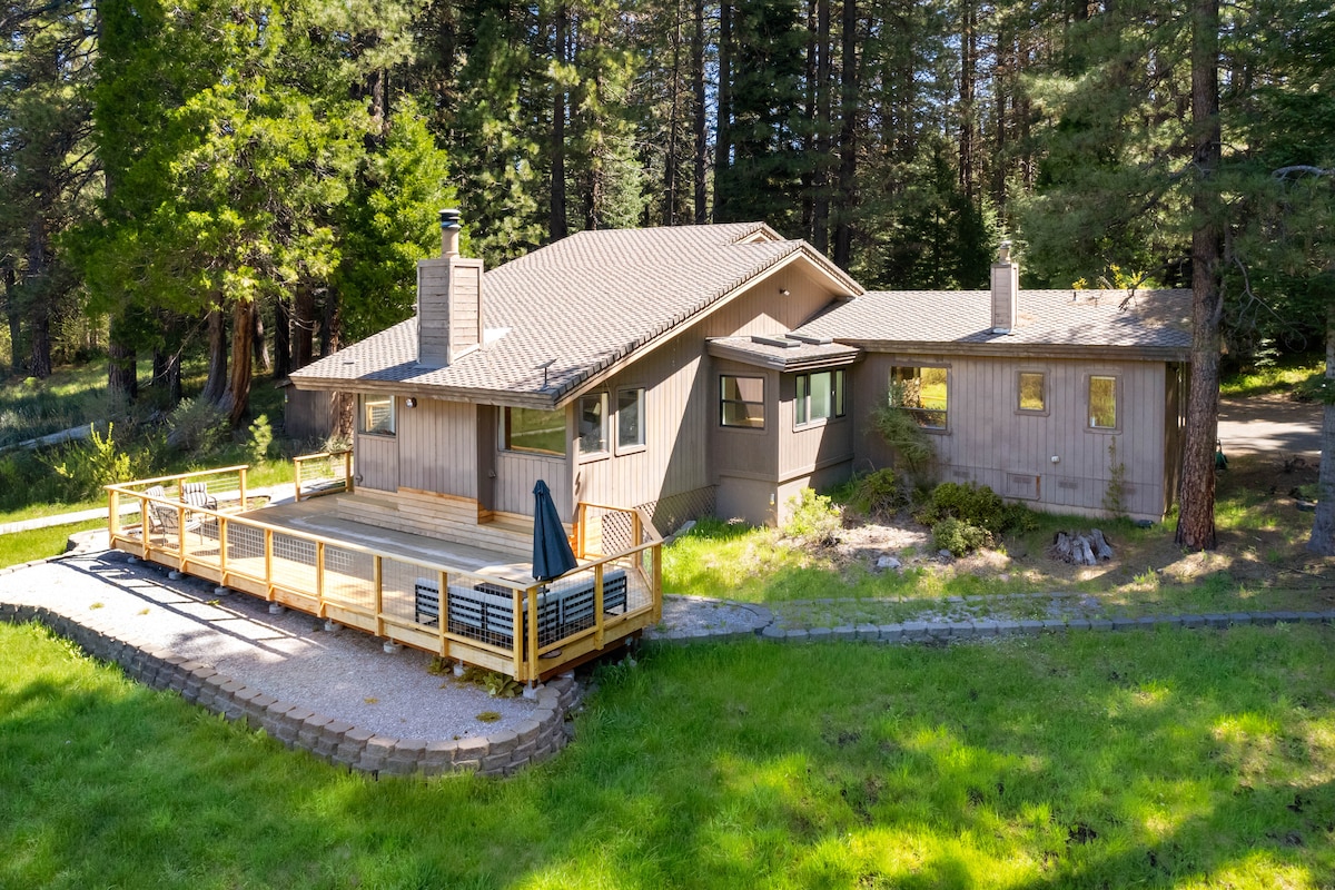 A spacious home is nestled among tall trees, featuring a wide wooden deck that encourages outdoor relaxation. The architecture combines modern elements with natural surroundings, while a well-kept yard offers a serene backdrop with lush greenery.