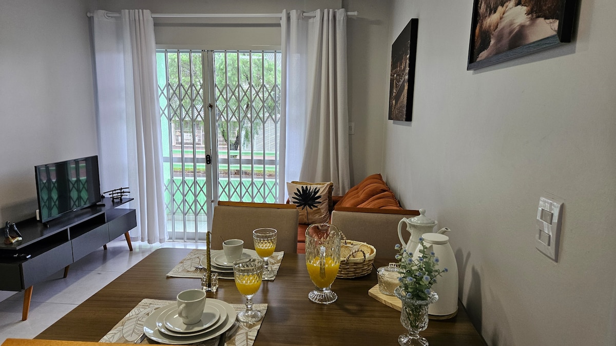 The dining area features a wooden table set for four, with plates and glasses arranged. A comfortable orange sofa is nearby, and a television stands on a low, modern console. Large glass doors lead to a green outdoor view, framed by light curtains.