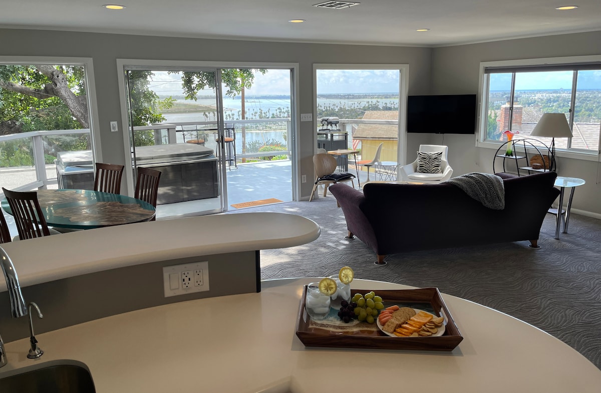 The open-concept living space features a dining area with wooden chairs and a round table, a comfortable couch facing expansive glass doors, and views of Mission Bay. A tray with sliced fruit and drinks rests on the kitchen counter, welcoming relaxation and enjoyment.