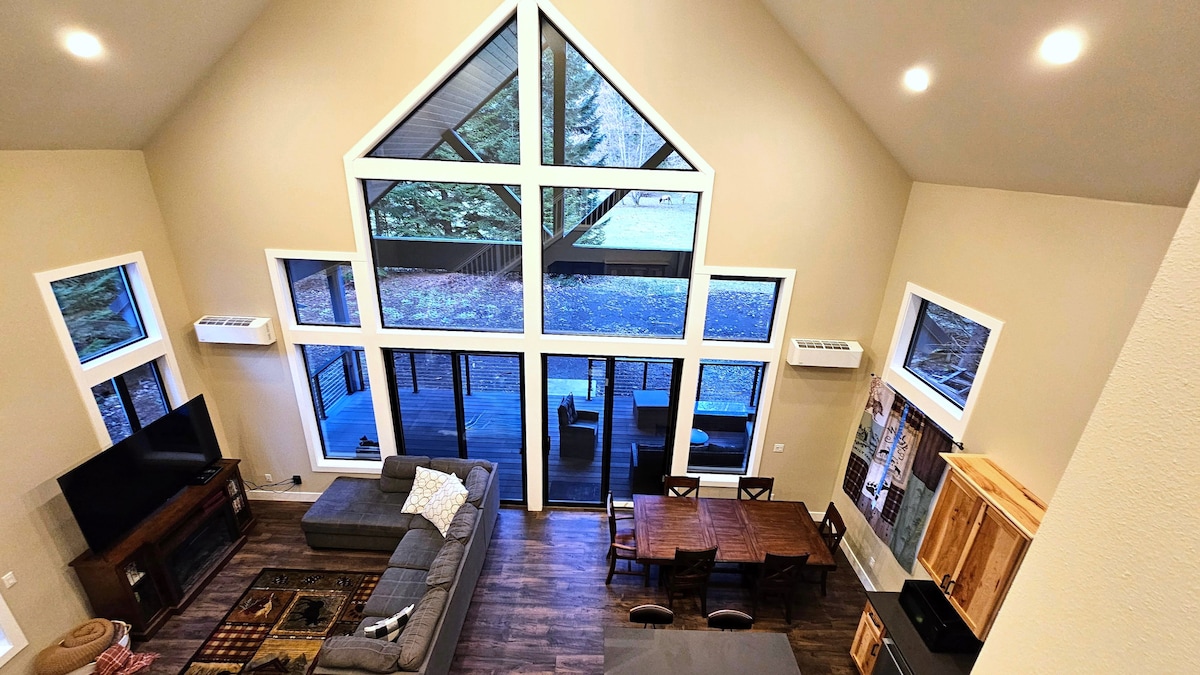A spacious living area displays vaulted ceilings with large picture windows offering views of the outdoor landscape. The open layout includes a comfortable sectional sofa, a large dining table, and a wall-mounted television, creating a welcoming environment for gatherings.
