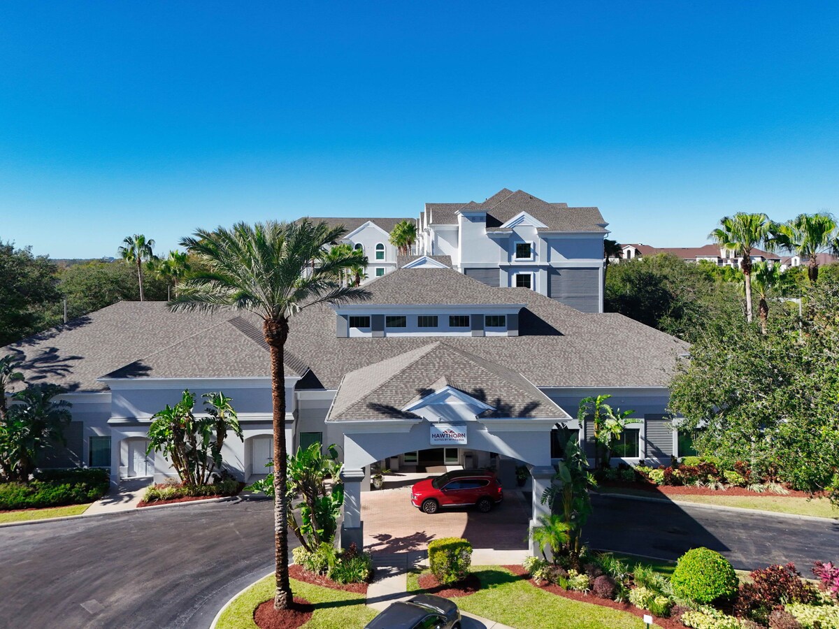The exterior of the Hawthorn Suites building is depicted, showcasing a well-maintained entrance. Lush greenery surrounds the area, complemented by palm trees. The parking lot is visible, with a red car parked under the porte-cochère, and blue skies provide a clear backdrop.