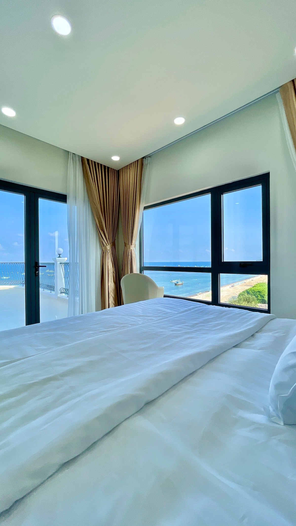 A spacious bedroom features large windows offering views of the beach and ocean. Soft lighting illuminates the room, while sheer curtains add a touch of elegance. A neatly made bed takes center stage, providing a comfortable space for relaxation.