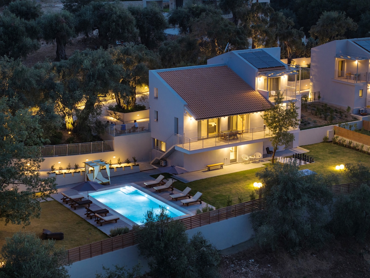 An aerial view captures multiple private villas surrounded by olive trees, showcasing an inviting outdoor area. The spacious gardens feature a heated pool, sun loungers, and comfortable seating. Soft lighting enhances the serene ambiance as evening sets in.