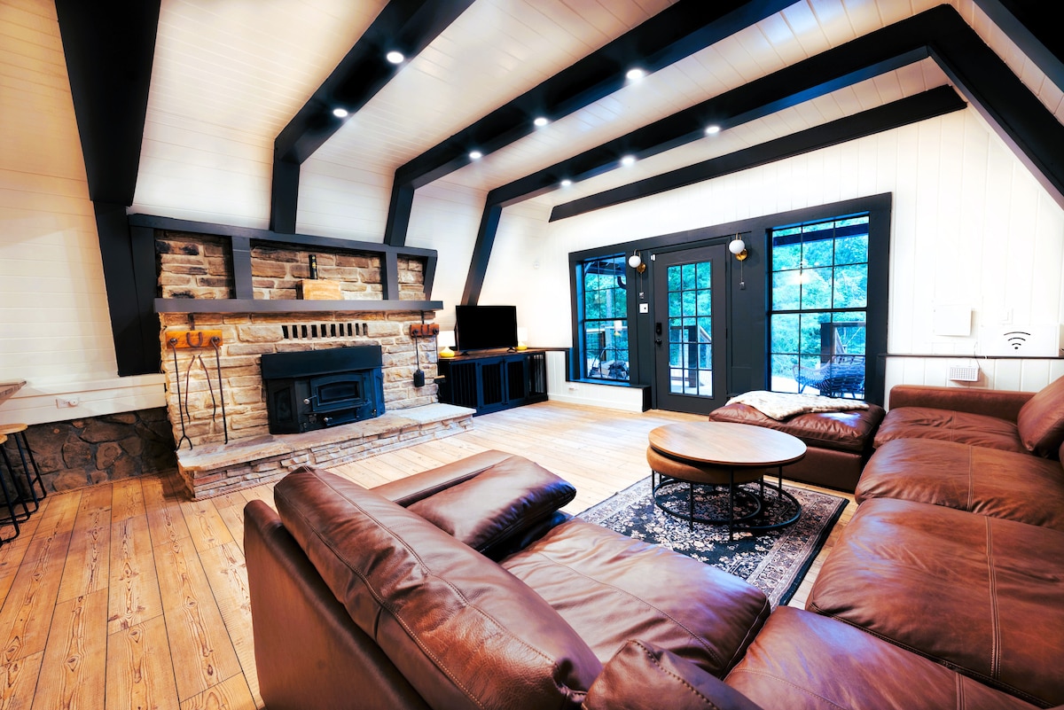 A spacious living area features a stone fireplace as a central focal point, complemented by dark wooden beams overhead. Comfortable leather seating is arranged around a circular table, with large glass doors providing views of the outdoors.