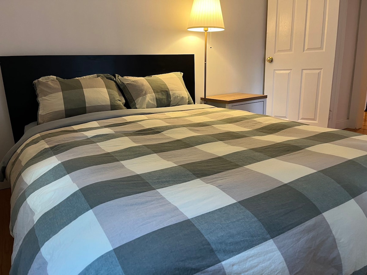 A spacious bed is dressed in a plaid duvet cover, featuring shades of grey and white. Soft light is provided by a standing lamp beside a minimalist bedside table. A closed white door is situated in the background.