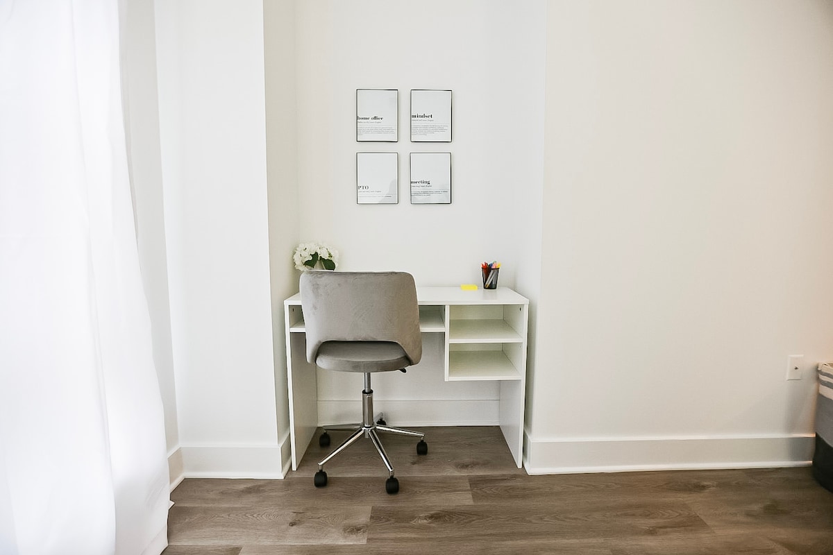 A designated office space is provided, featuring a clean, white desk with an ergonomic chair. Four framed prints adorn the wall above, and a small vase of flowers adds a touch of decor. Natural light filters in through a nearby window covered by sheer curtains.