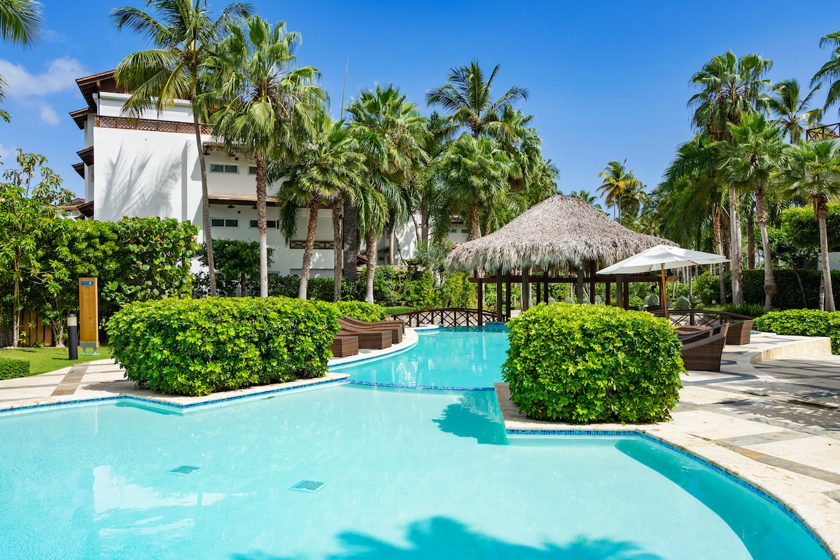A tranquil resort pool is surrounded by lush greenery and palm trees. A thatched roof gazebo provides shaded seating near the water. Clear blue skies reflect on the pool's surface, creating a serene atmosphere. Multiple lounge chairs are arranged around the pool for relaxation.