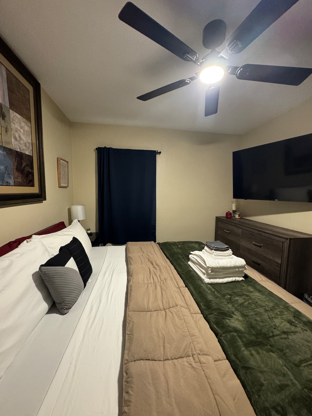 A spacious bedroom features a king bed topped with neatly arranged white towels. A dark-fan provides ceiling illumination, and a large flat-screen TV is mounted on the wall opposite the bed. A dark curtain covers the window, while artwork is displayed prominently.