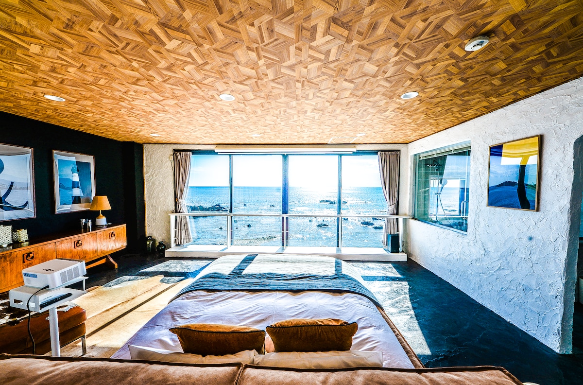 A spacious bedroom is presented with a wooden ceiling and large windows overlooking the ocean. The bed is positioned against a textured wall, complemented by two decorative pillows. Natural light floods the room, highlighting the sleek furnishings and inviting atmosphere.
