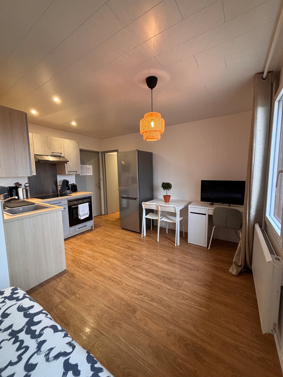 A modern kitchen area is visible, featuring light wood cabinetry and stainless steel appliances. A small dining table with two chairs is positioned beside a desk with a computer monitor. Natural light enters through a window draped with light fabric, while a decorative pendant light hangs from the ceiling.