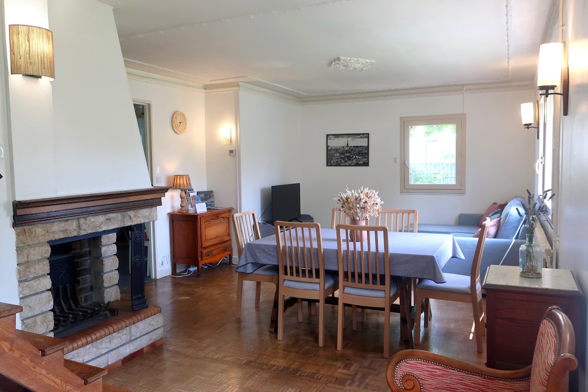 A spacious living and dining area is highlighted by natural light streaming through a window. A fireplace retains warmth, complementing a wooden dining table surrounded by chairs. A comfortable convertible sofa is positioned beside an entertainment unit with a television.
