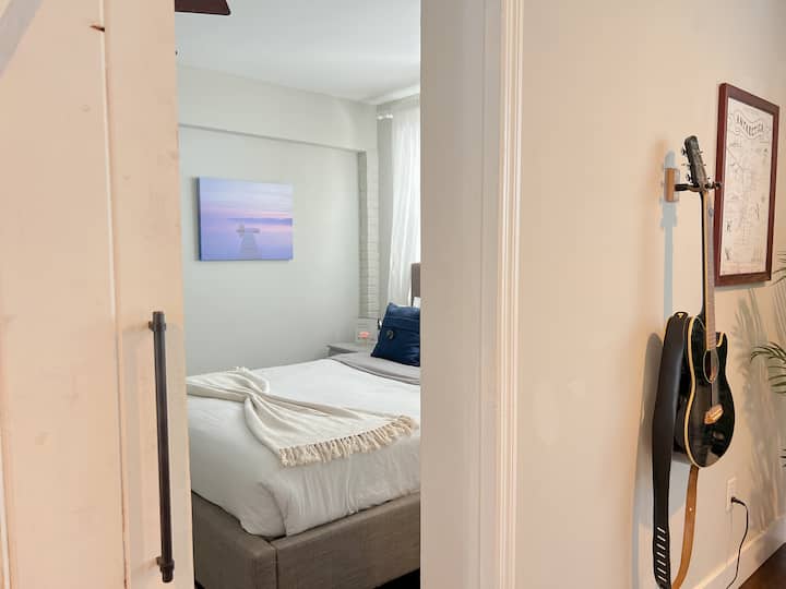 Sunrise calm & guitar charm — With beachy art, navy pillows, and a hanging axe, this bedroom blends restful vibes with a touch of rock & roll