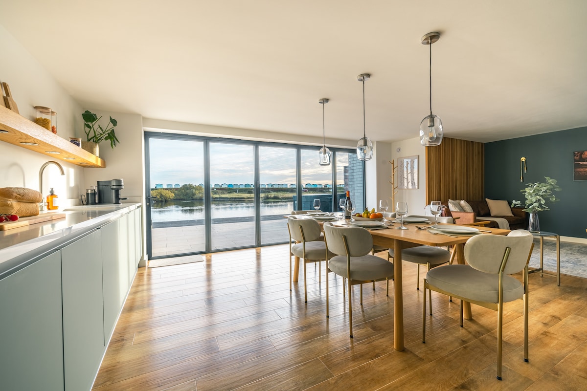 An open-plan kitchen and dining area features a long dining table surrounded by six chairs. Large glass doors provide a view of Widewater Lagoon, allowing natural light to fill the space. Minimalist cabinetry and warm wooden flooring add to the contemporary design.