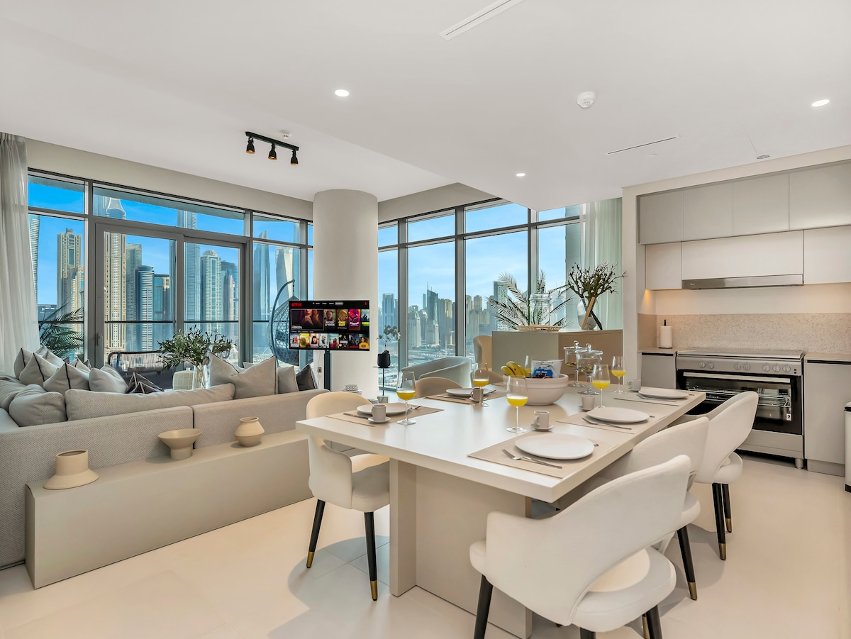 The modern kitchen and dining area feature a spacious table set for six, with elegant dishware arranged neatly. Natural light floods in through large windows, illuminating the sleek cabinetry and countertops, while a cozy seating area with a sofa is visible in the background.