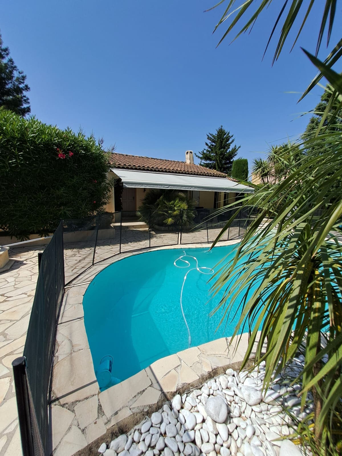A clear blue swimming pool is surrounded by a natural stone patio and a safety fence. Lush greenery, including palm trees, enhances the outdoor space, while a shaded area is located adjacent to the pool, providing a cool retreat from the sun.