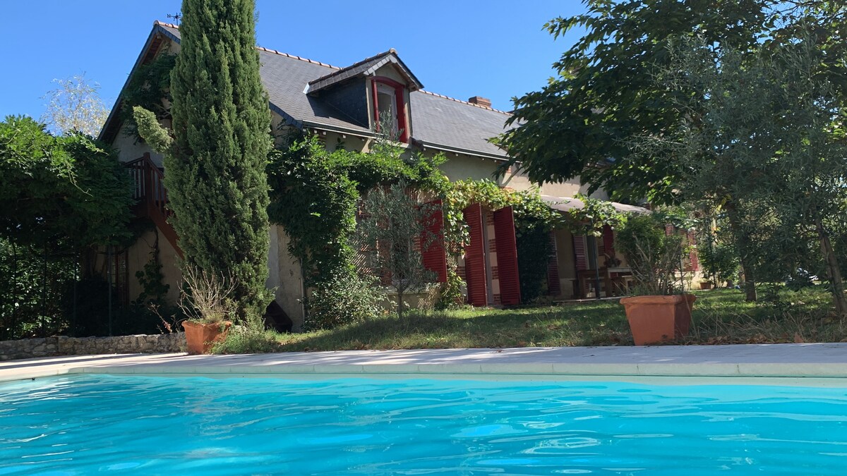 The exterior of the historic school building features rich greenery, with vibrant red shutters contrasting against the neutral facade. A private swimming pool is visible in the foreground, offering a refreshing blue invitation, while potted plants and sunlit garden spaces contribute to a serene outdoor setting.