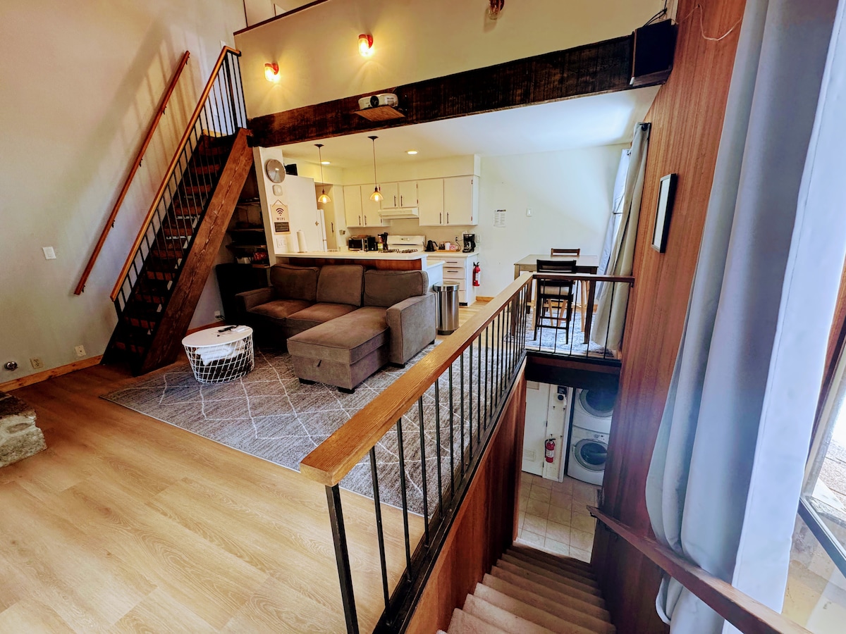 A spacious living area is visible, featuring a sectional sofa and a coffee table on a light gray rug. Stairs lead to the upper level, where a dining area and kitchen can be seen. Large windows allow natural light to illuminate the space.
