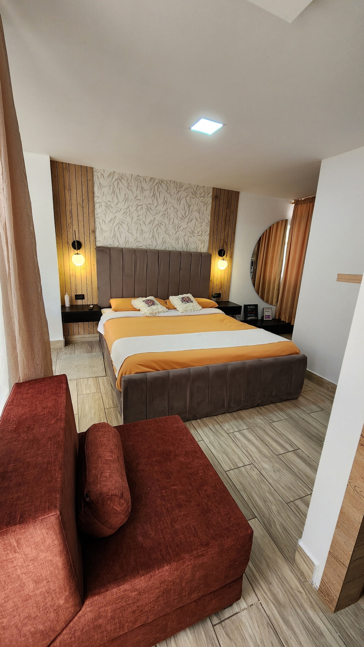 A spacious room features a queen bed with warm-toned bedding, complemented by decorative pillows. A sofa in rich orange sits in the corner. Soft natural light filters through sheer curtains, illuminating the light wood and textured wall accents.