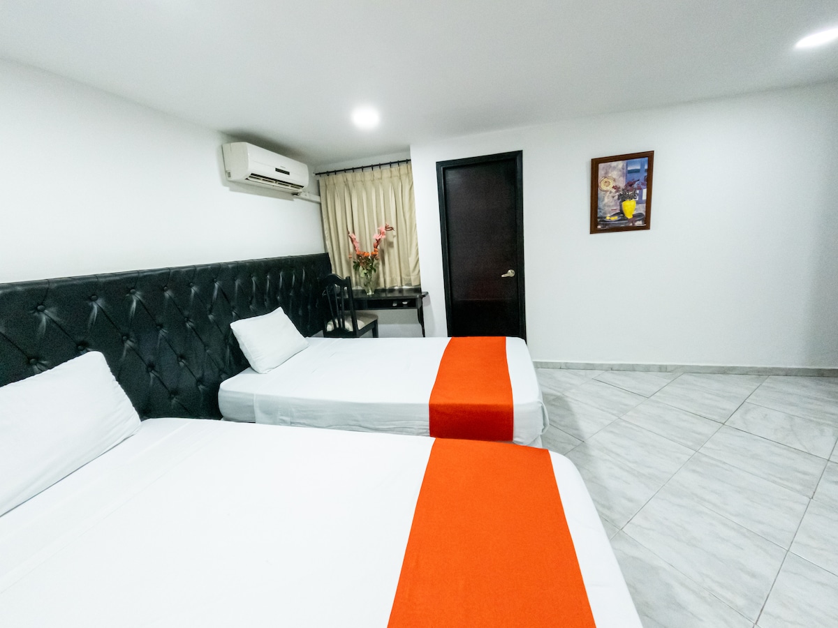 A well-appointed room features a spacious bed with crisp linens and a second smaller bed, both positioned against a stylish headboard. A decorative orange accent blanket adds a pop of color. An air conditioning unit is mounted on the wall, while soft lighting illuminates the space.