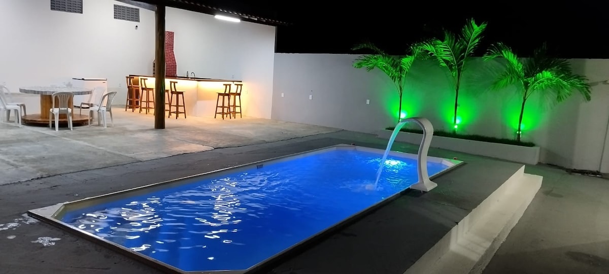 A clear blue pool is set against a minimalist outdoor area, illuminated by green lights. A dining table with several chairs and a bar area can be seen in the background, enhancing the gathering space for relaxation and entertainment.