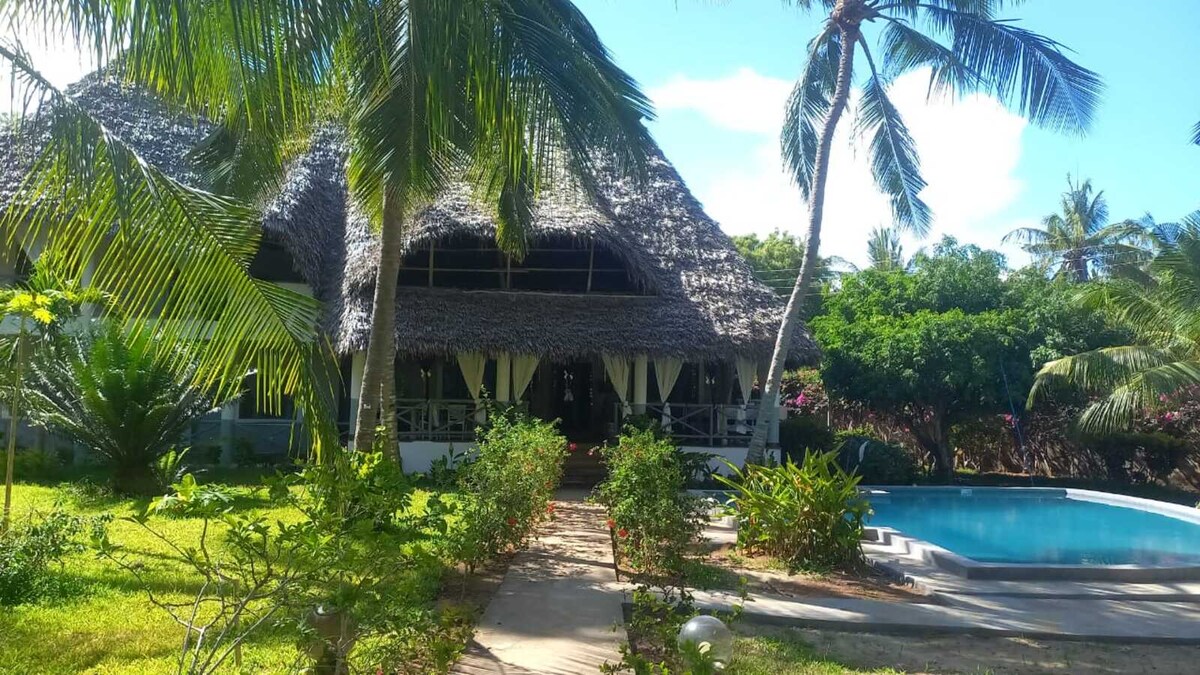 The villa features a traditional straw-thatched roof, nestled among lush palm trees. A pathway leads to the entrance, bordered by colorful tropical plants. A sparkling swimming pool is visible to the side, enhancing the serene outdoor setting.