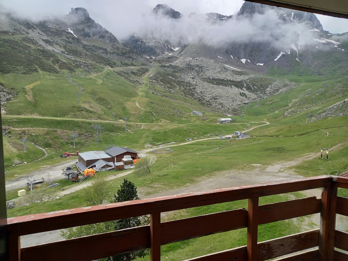 A scenic view of a mountainous landscape is presented from the balcony. Lush green hillsides are visible, with ski lifts and structures nestled in the valley below. Clouds gently envelop the mountain peaks, creating a serene backdrop for this tranquil setting.