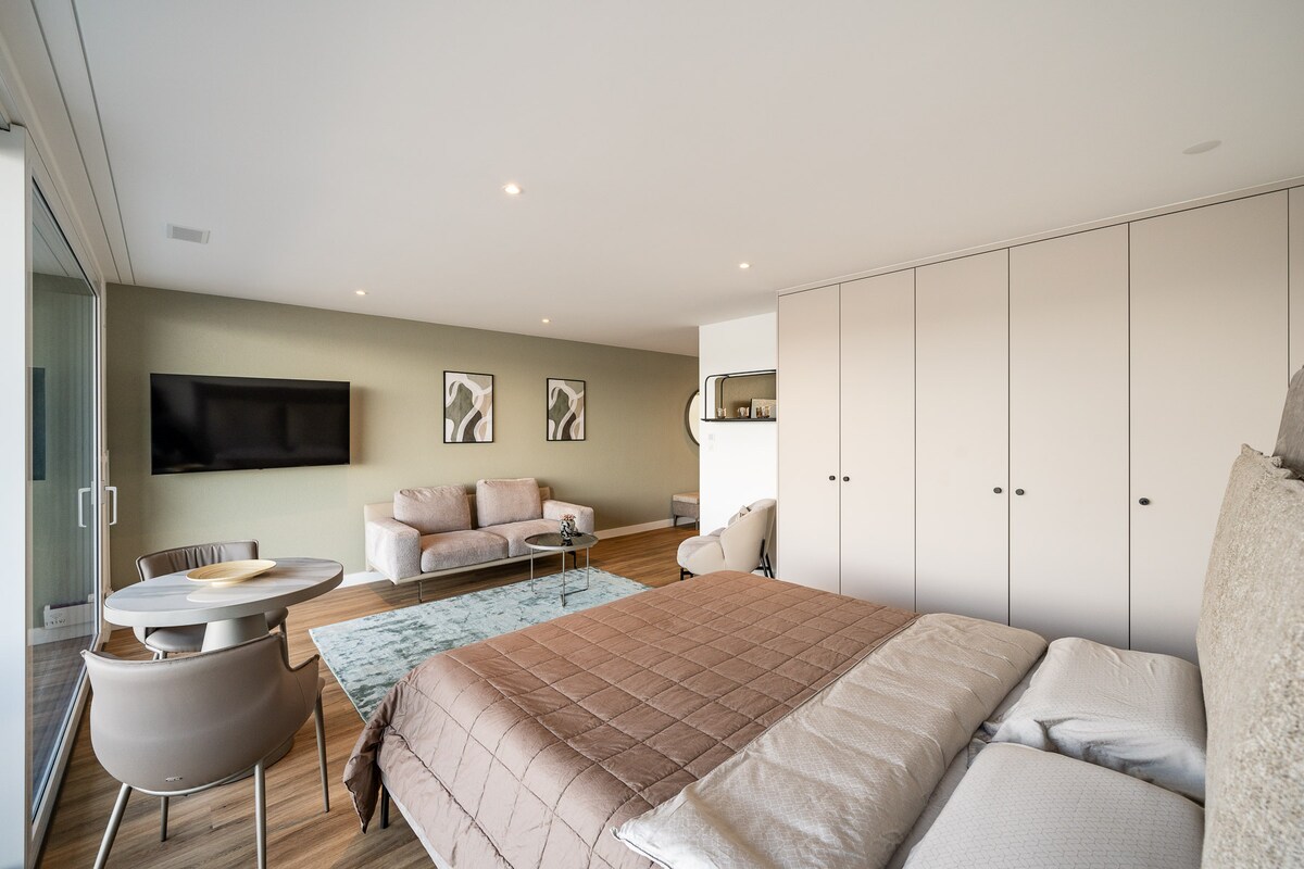 An open living area is visible, featuring a spacious bed with neutral bedding. A soft area rug lies beneath a round dining table and four chairs. A comfortable seating arrangement is present, complemented by wall art and a television mounted on a light green wall.