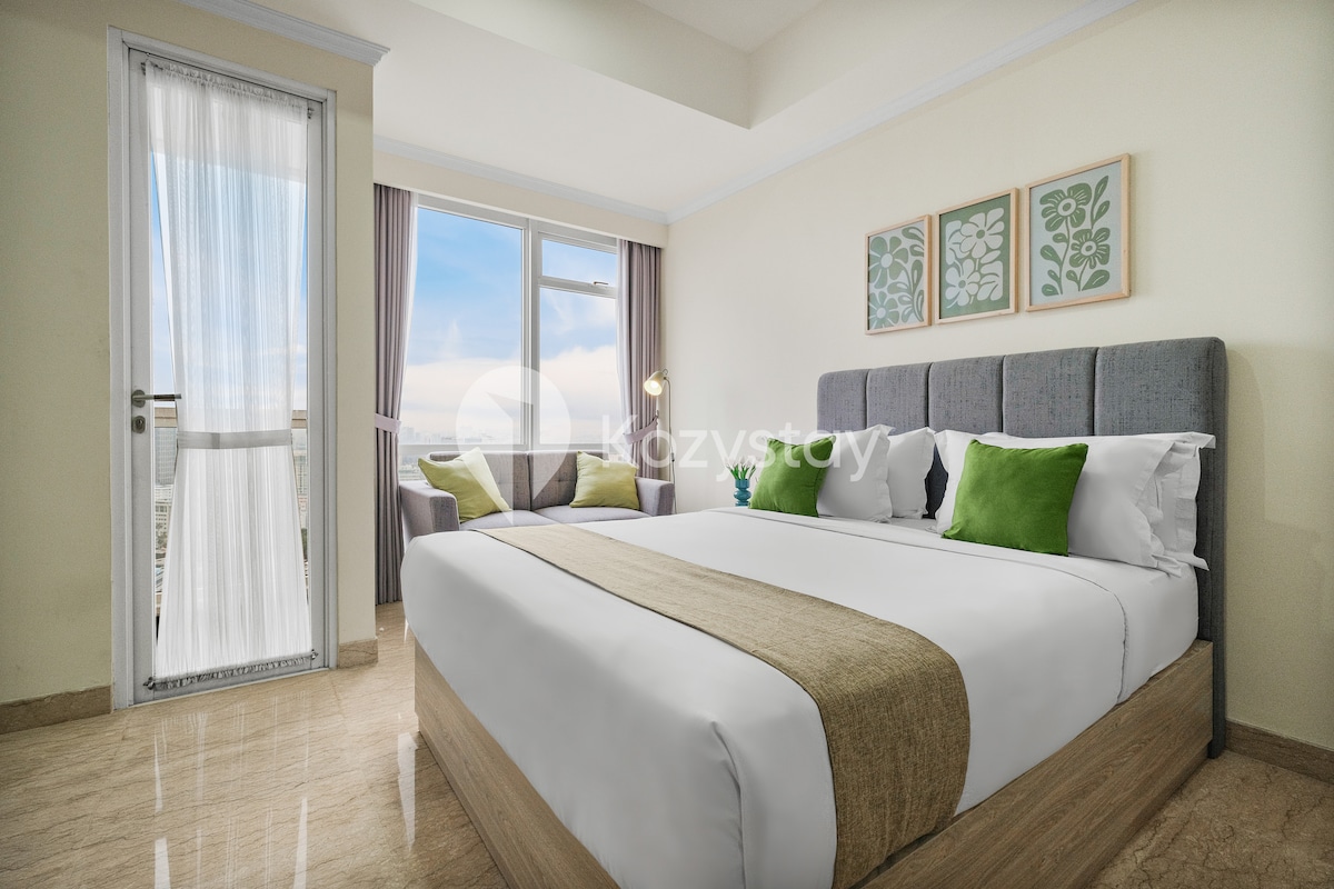 A bright bedroom features a spacious bed adorned with white linens and an earthy-toned runner. Two green accent pillows complement the neutral headboard. Natural light floods in from the large window, which is dressed with sheer curtains, and a private balcony is accessible.